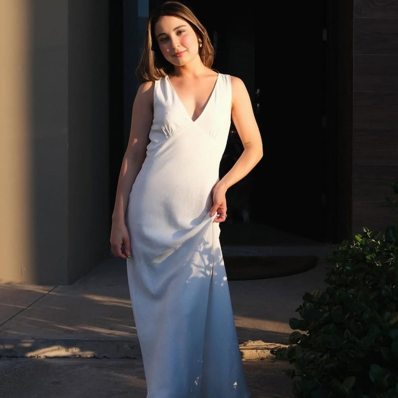 May include: A white, sleeveless maxi dress with a V-neck. The dress is floor-length and appears to be made of a lightweight fabric. The model is standing outside in front of a dark doorway.