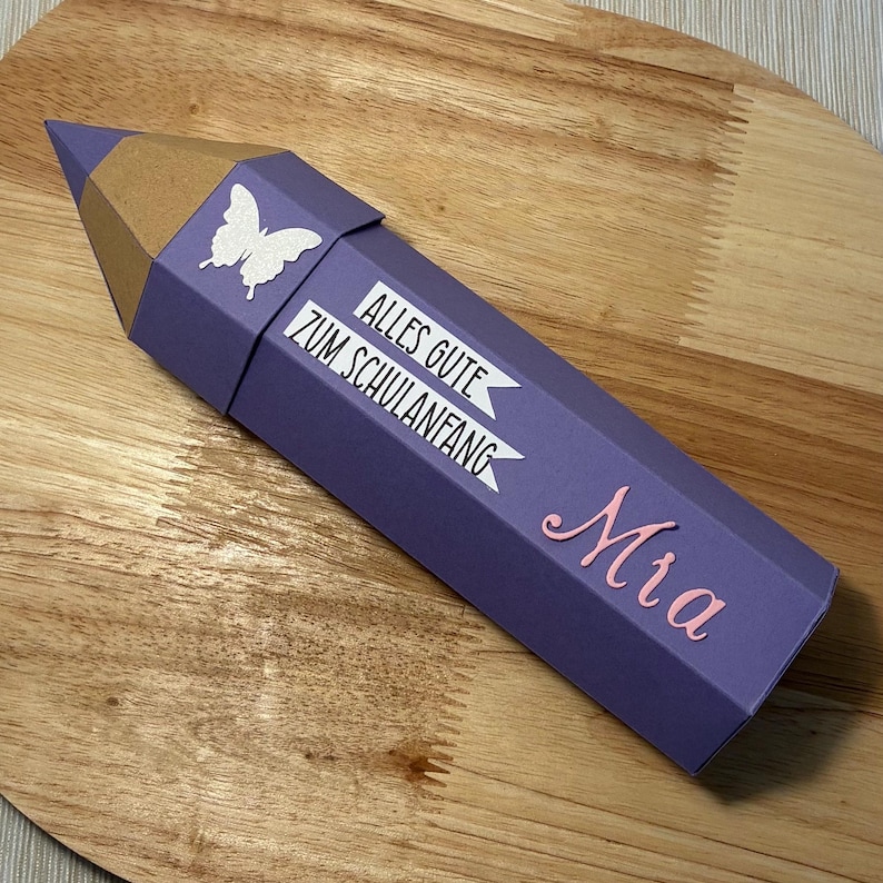 Pode incluir: Uma caixa de presente em forma de l&aacute;pis roxo com ponta marrom. A caixa tem uma borboleta branca e o texto "ALLES GUTE ZUM SCHULANFANG". O nome "Mia" est&aacute; escrito em rosa. Em uma superf&iacute;cie de madeira.