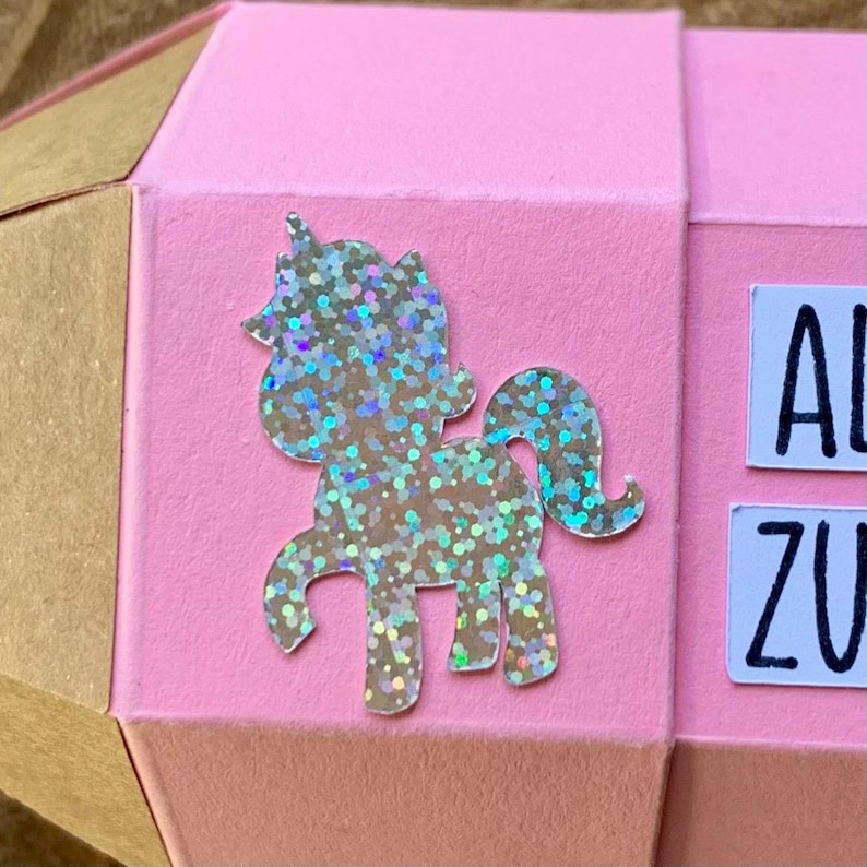 Pode incluir: Um artesanato em papel rosa e geom&eacute;trico com um adesivo de unic&oacute;rnio brilhante. O unic&oacute;rnio &eacute; prateado com detalhes iridescentes. O artesanato tem um esquema de cores marrom e rosa, com as letras "AL" e "ZU" vis&iacute;veis.