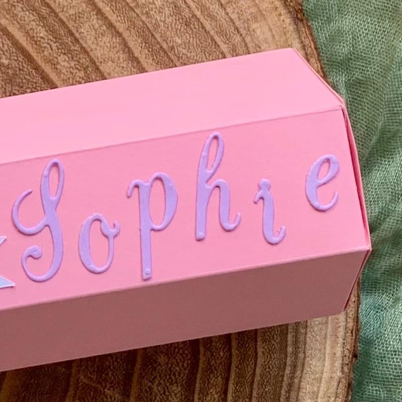 Pode incluir: Uma caixa de presente rosa com o nome "Sophie" em escrita lavanda. A caixa tem formato hexagonal e est&aacute; sobre uma superf&iacute;cie de madeira e um tecido verde.