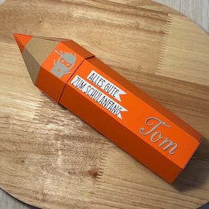 Pode incluir: Uma caixa de presente laranja em forma de l&aacute;pis com ponta castanha. A caixa tem o texto "Alles Gute zum Schulanfang" e o nome "Tom" em prata. Um gr&aacute;fico ninja tamb&eacute;m &eacute; vis&iacute;vel.