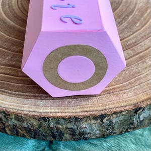 Pode incluir: Uma caixa de presente rosa em forma de l&aacute;pis hexagonal com um c&iacute;rculo castanho na extremidade. A parte superior da caixa tem a palavra "be" em it&aacute;lico, em uma fonte roxa clara. A caixa est&aacute; em uma superf&iacute;cie de madeira.