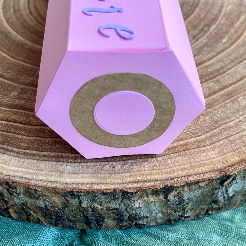 Pode incluir: Uma caixa de presente rosa em forma de l&aacute;pis hexagonal com um c&iacute;rculo castanho na extremidade. A parte superior da caixa tem a palavra "be" em it&aacute;lico, em uma fonte roxa clara. A caixa est&aacute; em uma superf&iacute;cie de madeira.