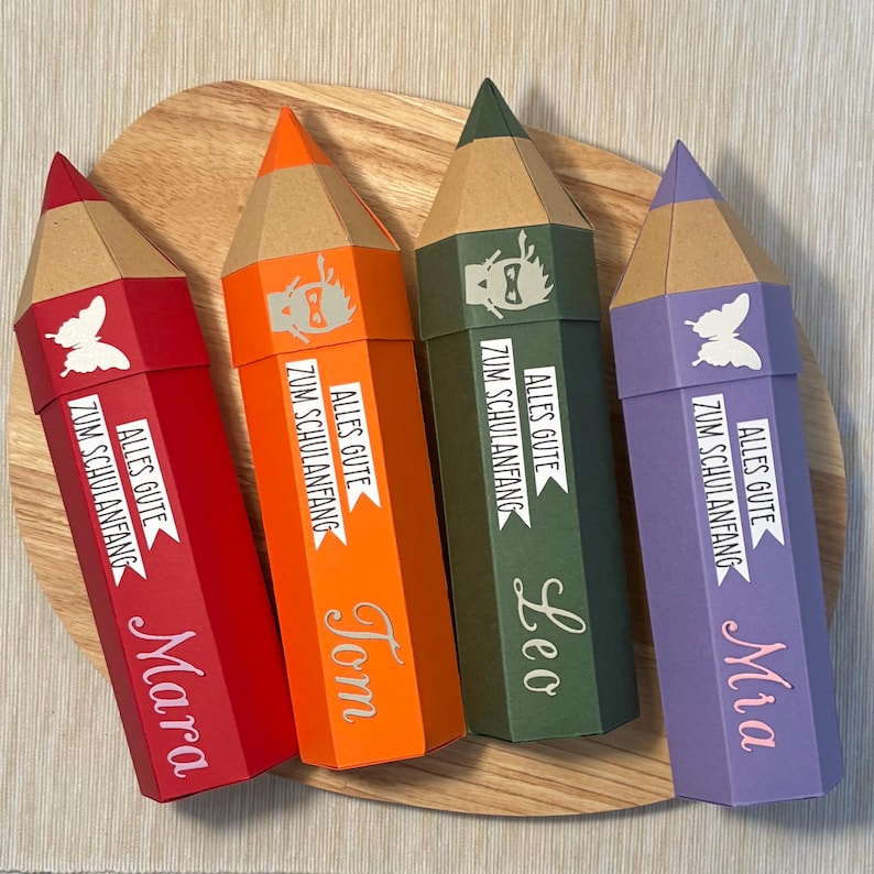 Pode incluir: Quatro caixas de presente coloridas em forma de l&aacute;pis. As caixas s&atilde;o vermelhas, laranja, verdes e roxas. Cada caixa tem um nome, um gr&aacute;fico de borboleta ou super-her&oacute;i e o texto "Alles Gute Zum Schulanfang". Em uma superf&iacute;cie de madeira.