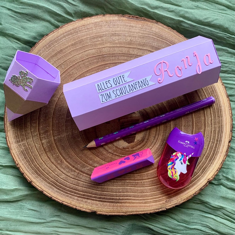 Pode incluir: Um conjunto de material escolar, incluindo um estojo roxo com o nome "Ronja", um l&aacute;pis, uma borracha e um apontador com um design de unic&oacute;rnio. O estojo tem o texto "ALLES GUTE ZUM SCHULANFANG".
