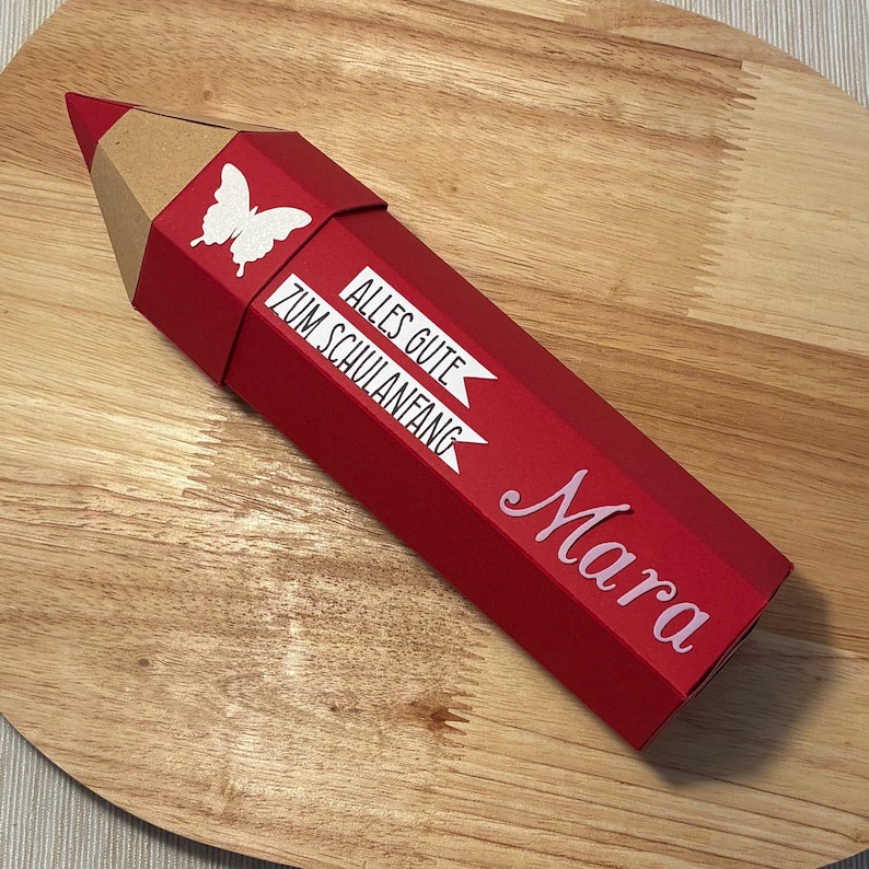 Pode incluir: Uma caixa de presente vermelha em forma de l&aacute;pis com uma borboleta e o texto "Alles Gute Zum Schulanfang" e o nome "Mara" em escrita branca. A ponta do l&aacute;pis &eacute; castanha.