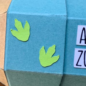Pode incluir: Close-up de um artesanato geom&eacute;trico em papel azul claro com dois recortes de pegadas de dinossauro verde lim&atilde;o. Azulejos de letras brancas soletram "A" e "Z". O artesanato em papel tem um lado castanho e dourado.