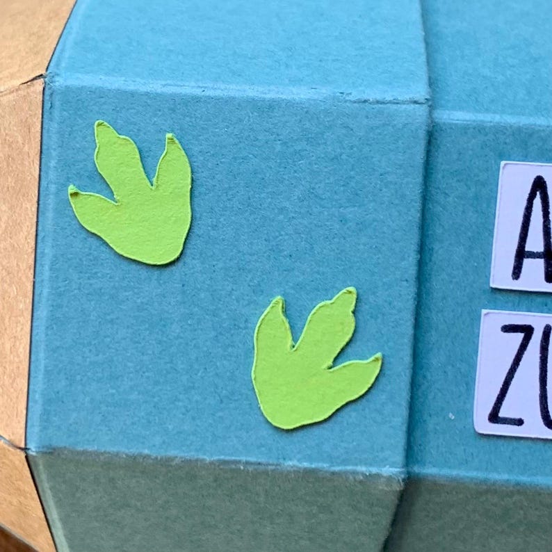 Pode incluir: Close-up de um artesanato geom&eacute;trico em papel azul claro com dois recortes de pegadas de dinossauro verde lim&atilde;o. Azulejos de letras brancas soletram "A" e "Z". O artesanato em papel tem um lado castanho e dourado.