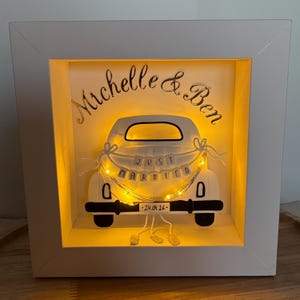 Könnte beinhalten: Weiß gerahmte Schattenbox mit weißem Auto-Ausschnitt, Lichterkette und den Worten "Just Married". Die Namen "Michelle & Ben" stehen über dem Auto. Das Datum "24.04.26" steht darunter.