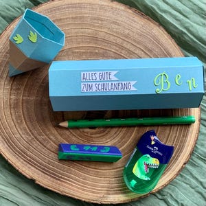 Pode incluir: Uma caixa azul clara em forma de l&aacute;pis com o texto "Alles Gute zum Schulanfang" e o nome "Ben". Tamb&eacute;m est&atilde;o presentes um l&aacute;pis verde, uma borracha e um apontador com um gr&aacute;fico de dinossauro. Os itens est&atilde;o em uma superf&iacute;cie de madeira.