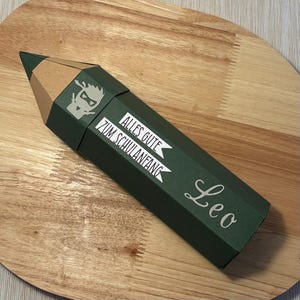 Pode incluir: Uma caixa de presente em forma de l&aacute;pis verde escuro com uma ponta castanha. A caixa tem texto branco que diz "ALLES GUTE ZUM SCHULANFANG" e o nome "Leo". Um gr&aacute;fico de um ninja de desenho animado tamb&eacute;m &eacute; vis&iacute;vel.