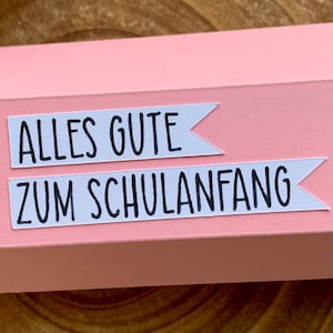 Pode incluir: Uma caixa de presente rosa claro com duas etiquetas brancas. As etiquetas, em forma de banner, exibem o texto "ALLES GUTE" e "ZUM SCHULANFANG" em preto. A caixa est&aacute; sobre uma superf&iacute;cie de madeira.