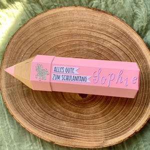 Pode incluir: Uma caixa de presente rosa em forma de l&aacute;pis com ponta dourada. A caixa tem o texto "Alles Gute Zum Schulanfang" e o nome "Sophie" em it&aacute;lico. Um unic&oacute;rnio prateado com glitter tamb&eacute;m est&aacute; na caixa. A caixa est&aacute; sobre uma superf&iacute;cie de madeira.