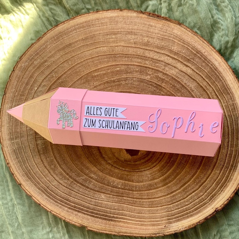 Pode incluir: Uma caixa de presente rosa em forma de l&aacute;pis com ponta dourada. A caixa tem o texto "Alles Gute Zum Schulanfang" e o nome "Sophie" em it&aacute;lico. Um unic&oacute;rnio prateado com glitter tamb&eacute;m est&aacute; na caixa. A caixa est&aacute; sobre uma superf&iacute;cie de madeira.
