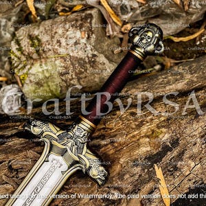 Pode incluir: Uma espada decorativa com um pomo em forma de caveira e uma guarda dourada ornamentada. A lâmina é prateada com detalhes gravados. O cabo é envolto em um material vermelho escuro. A espada repousa sobre uma superfície de madeira envelhecida.