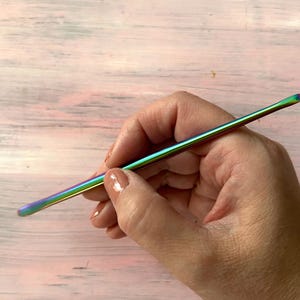 May include: A rainbow iridescent metal crochet hook held in a hand.