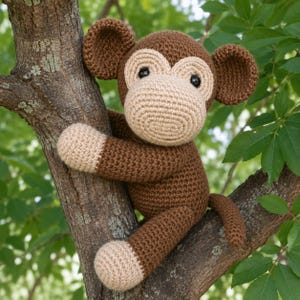 Op de afbeelding: Een handgemaakte gehaakte aap in een boom. De aap is bruin met beige accenten op zijn gezicht, handen en voeten. Hij heeft zwarte ogen en grote, ronde oren. De aap omhelst een tak, met groene bladeren op de achtergrond.