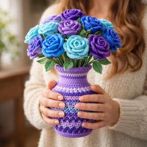 Può includere: Un vaso all'uncinetto pieno di un bouquet di rose fatte a mano. Le rose sono in tonalità di blu e viola, con foglie verdi. Il vaso è viola con un motivo decorativo. Un'arrangiamento floreale unico e artigianale.