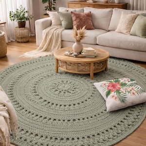 Könnte beinhalten: Ein runder, salbeigrüner Häkelteppich dominiert den Vordergrund eines Wohnzimmers. Der Teppich, mit einem Durchmesser von etwa 200 cm, zeigt konzentrische Kreismuster und liegt auf einem Holzboden. Ein Blumenkissen und ein Sofa sind ebenfalls zu sehen.