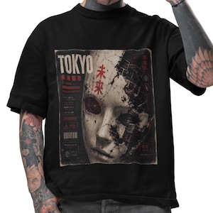 以下が含まれることがあります： 黒いTシャツ。赤目とロボット要素を持つ、顔のグラフィックが描かれています。グラフィックには日本語のテキストと「TOKYO」の文字が含まれています。Tシャツはクルーネックで半袖です。