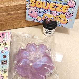 Könnte beinhalten: Ein lilafarbenes, weiches Spielzeug in Form einer Blume mit einem Schmetterling in der Mitte und perlmuttartigen Akzenten. Das Spielzeug ist in einer durchsichtigen Plastiktüte verpackt. Der Hintergrund zeigt ein Schild mit der Aufschrift "Stress Free Squeeze" und Comicfiguren.