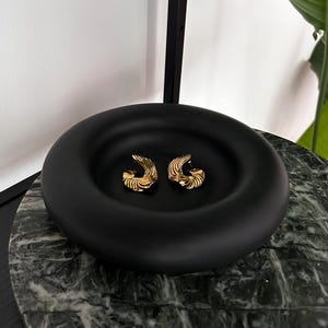 Könnte beinhalten: Ein Paar goldfarbene, strukturierte Creolen in einer schwarzen, runden Schale auf einer grünen Marmorfläche.