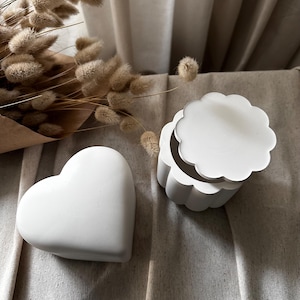 May include: Two white ceramic trinket boxes, one shaped like a heart and the other with a scalloped lid. The boxes are sitting on a light brown fabric surface.