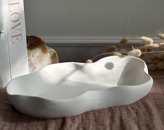 May include: A white, organically shaped ceramic bowl. The bowl has a smooth, matte finish and a shallow, elongated design. The bowl is displayed on a brown surface, with a book that says "LOVE" in the background.