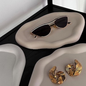 Könnte beinhalten: Zwei weiße Keramikteller mit einer gold umrandeten Sonnenbrille auf einem Teller und einem Paar goldenen Ohrringen auf dem anderen Teller.