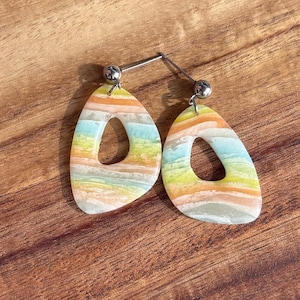 May include: A pair of colorful, abstract earrings. Each earring is a teardrop shape with a large, open center. The earrings feature horizontal stripes of pastel colors, including orange, yellow, blue, and white. The earrings are attached to silver posts.
