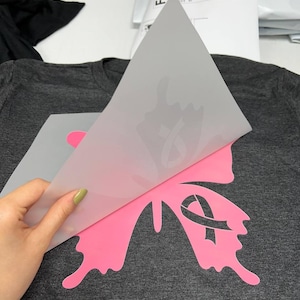 K&ouml;nnte beinhalten: Ein dunkelgraues T-Shirt mit einem rosa Schmetterlingsdesign mit einem rosa Band. Das Design wird mit einem W&auml;rme&uuml;bertragungsverfahren auf das Shirt aufgebracht. Das Schmetterlingsdesign ist ein Symbol f&uuml;r Brustkrebs-Aufkl&auml;rung.