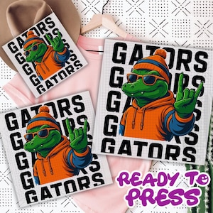 May include: A graphic design featuring a green alligator wearing sunglasses, an orange hoodie, and a blue and orange beanie, making a hand gesture. The word "GATORS" is printed around the alligator. The phrase "READY TO PRESS" is also visible.