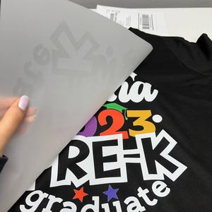 K&ouml;nnte beinhalten: Schwarzes T-Shirt mit einem farbenfrohen "Pre-K graduate 2023" Design. Das Design beinhaltet eine Abschlusskappe, Sterne und die Worte "Pre-K graduate" in fetter, wei&szlig;er Schrift. Eine durchscheinende Folie wird abgezogen.
