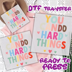 May include: DTF transfer sheets with the text "YOU CAN DO HARD THINGS" in colorful letters. The design includes hearts and is ready to press. The sheets are on a grid background.