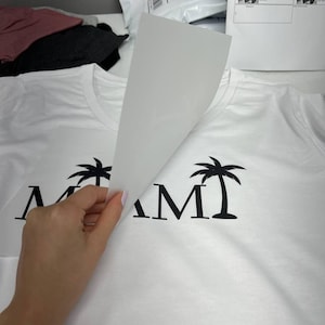 K&ouml;nnte beinhalten: Wei&szlig;es T-Shirt mit schwarzem "MIAMI"-Aufdruck mit Palmen. Das Design wird durch Anheben eines wei&szlig;en Blattes enth&uuml;llt. Andere Hemden und Versandmaterialien befinden sich im Hintergrund.