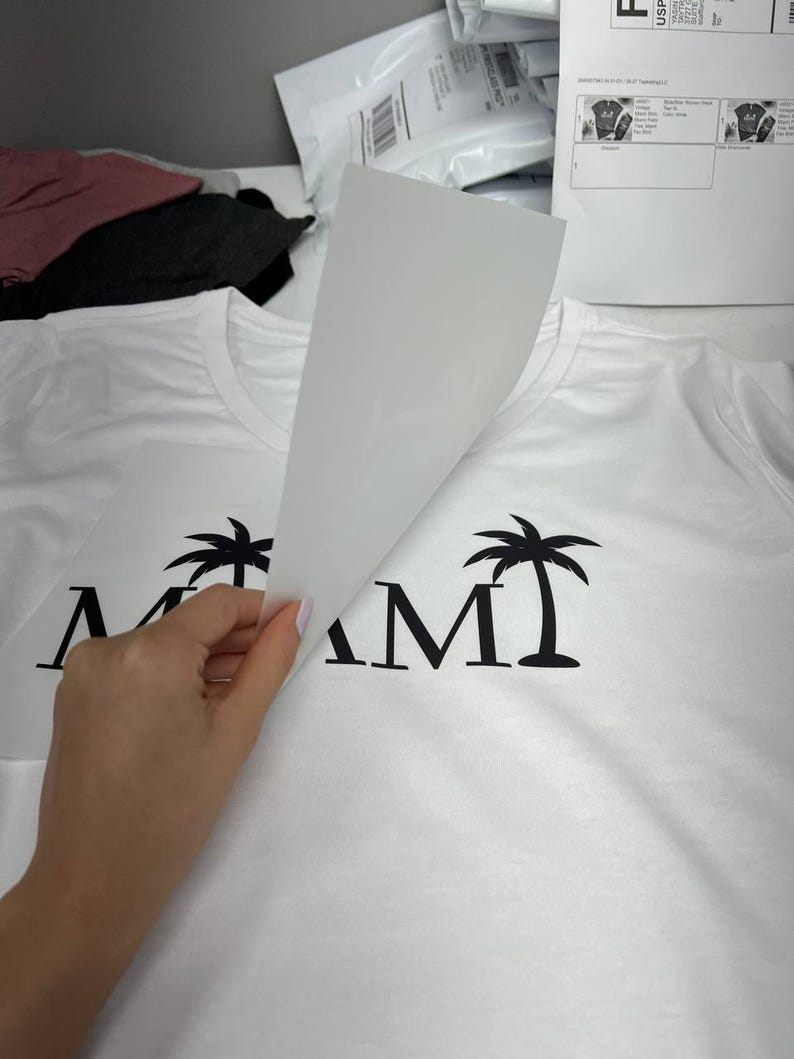 K&ouml;nnte beinhalten: Wei&szlig;es T-Shirt mit schwarzem "MIAMI"-Aufdruck mit Palmen. Das Design wird durch Anheben eines wei&szlig;en Blattes enth&uuml;llt. Andere Hemden und Versandmaterialien befinden sich im Hintergrund.