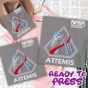 May include: Gray ready-to-press transfer sheets with the NASA Artemis logo, featuring a stylized moon and Earth design with red and blue accents. The text "READY TO PRESS" is in purple. A pink t-shirt and a brown hat are in the background.