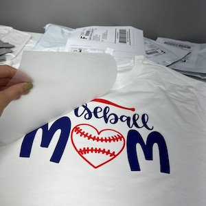 K&ouml;nnte beinhalten: Wei&szlig;es T-Shirt mit dem Schriftzug "baseball mom" in Marineblau und Rot. Das Wort "baseball" ist kursiv geschrieben, und das "o" in "mom" ist durch ein rotes Baseballherz ersetzt.