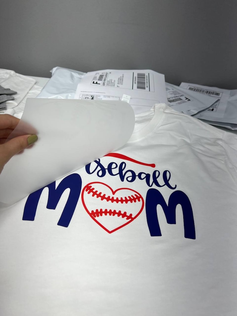 K&ouml;nnte beinhalten: Wei&szlig;es T-Shirt mit dem Schriftzug "baseball mom" in Marineblau und Rot. Das Wort "baseball" ist kursiv geschrieben, und das "o" in "mom" ist durch ein rotes Baseballherz ersetzt.