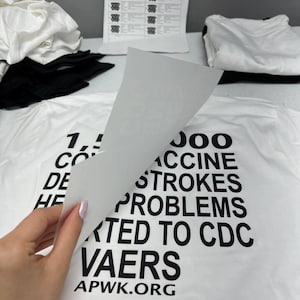 K&ouml;nnte beinhalten: Ein wei&szlig;es T-Shirt mit schwarzem Text, der lautet: "1.000.000 IMPFST&Ouml;SSE PROBLEME GEMELDET AN CDC VAERS APWK.ORG". Eine Hand hebt eine Schicht des Shirts an. Weitere wei&szlig;e und schwarze Shirts sind im Hintergrund.