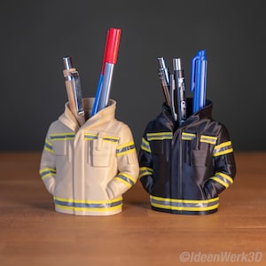 Könnte beinhalten: Zwei Stifthalter in Form von Feuerwehrjacken. Einer ist beige mit gelben und grauen Streifen, der andere ist schwarz mit gelben und grauen Streifen. Stifte und Marker sind in den Haltern sichtbar. Die Stifthalter stehen auf einer Holzoberfläche.