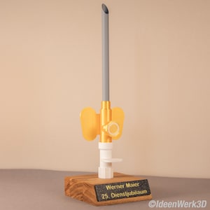 Könnte beinhalten: Ein 3D-gedrucktes medizinisches Gerät mit einem gelben schmetterlingsförmigen Element, einem grauen Rohr und einer weißen Basis, montiert auf einem Holzblock. Der Block hat eine schwarze Plakette mit dem Text "Werner Maier 25. Dienstjubiläum".
