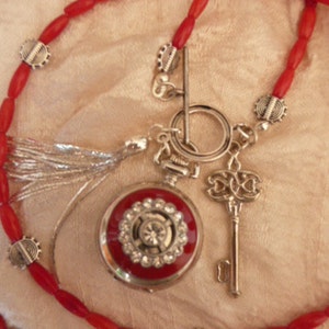 May include: A silver pocket watch with a red face and a white dial is attached to a red beaded necklace with silver accents. The watch has a key attached to it.