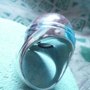 Pu&ograve; includere: Un anello d'argento con una pietra preziosa blu incastonata al centro. L'anello ha una fascia larga e una finitura liscia e lucida.