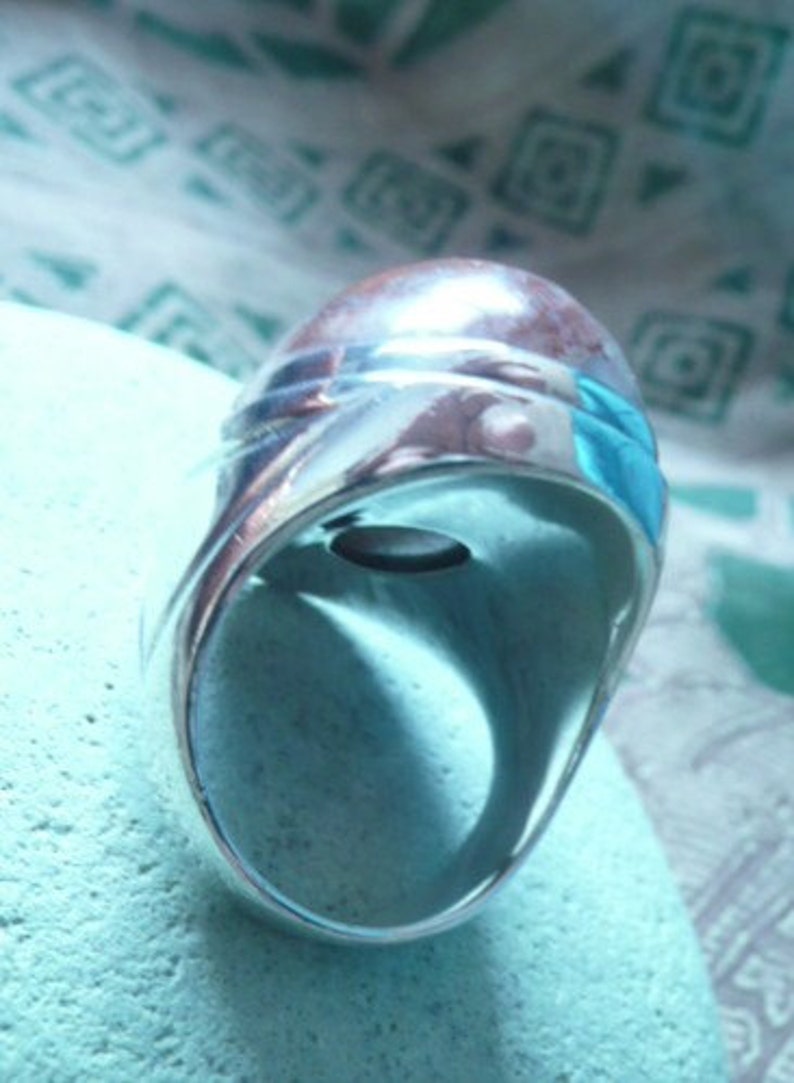 Pu&ograve; includere: Un anello d'argento con una pietra preziosa blu incastonata al centro. L'anello ha una fascia larga e una finitura liscia e lucida.
