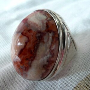 Puede incluir: Un anillo de plata con una piedra grande de forma ovalada. La piedra es una mezcla de colores blancos, rosados y marrones, con un patrón de remolino.