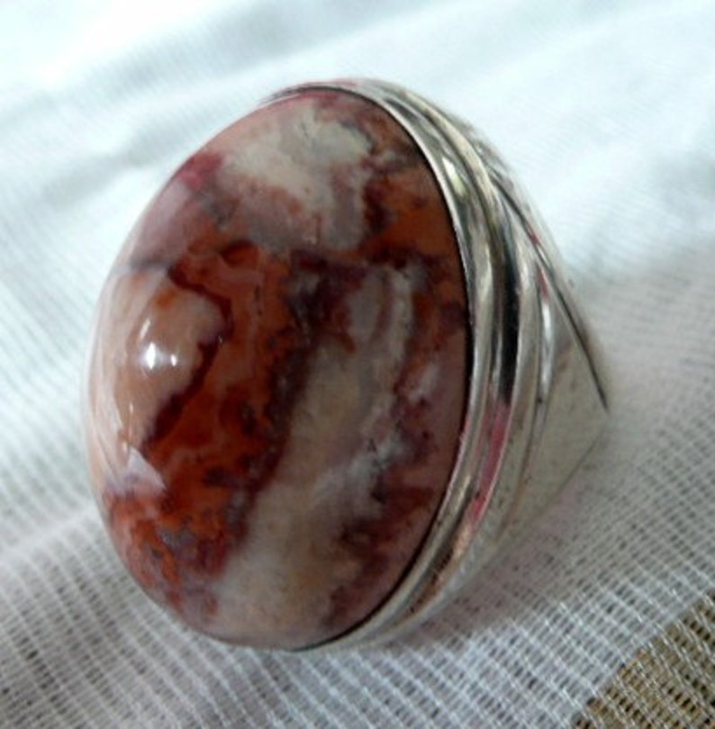 Pu&ograve; includere: Un anello d'argento con una grande pietra ovale. La pietra &egrave; un mix di colori bianchi, rosa e marroni, con un motivo a spirale.