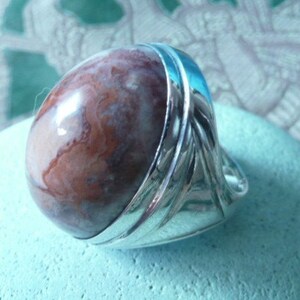 Pu&ograve; includere: Un anello d'argento con una grande pietra di agata rotonda, di colore marrone e bianco, incastonata al centro. L'anello ha una fascia testurizzata con un design decorativo.