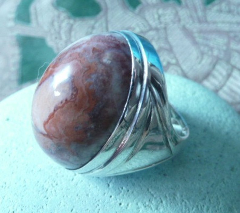 Pu&ograve; includere: Un anello d'argento con una grande pietra di agata rotonda, di colore marrone e bianco, incastonata al centro. L'anello ha una fascia testurizzata con un design decorativo.