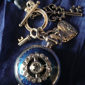 May include: A silver necklace with a blue lapis lazuli stone pendant, a key charm, a heart charm with the inscription "Love you more", and a pocket watch with a blue face and rhinestones.